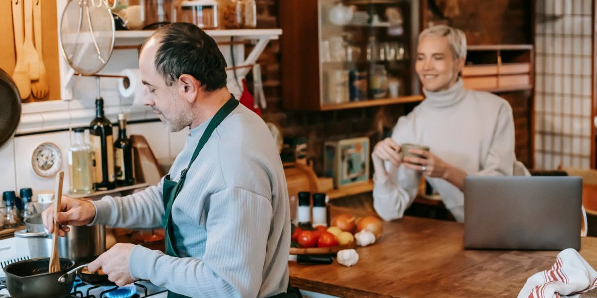 Focused male preparing dish on gas stove in kitchen with smiling wife having drink and using laptop