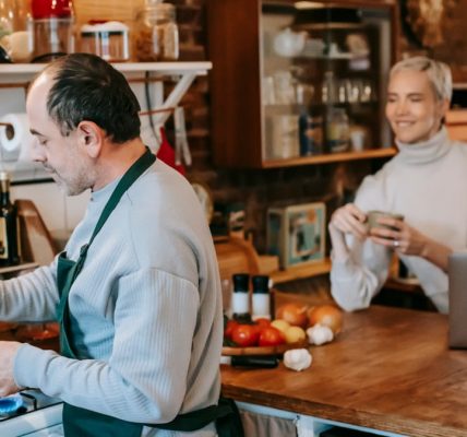 Focused male preparing dish on gas stove in kitchen with smiling wife having drink and using laptop
