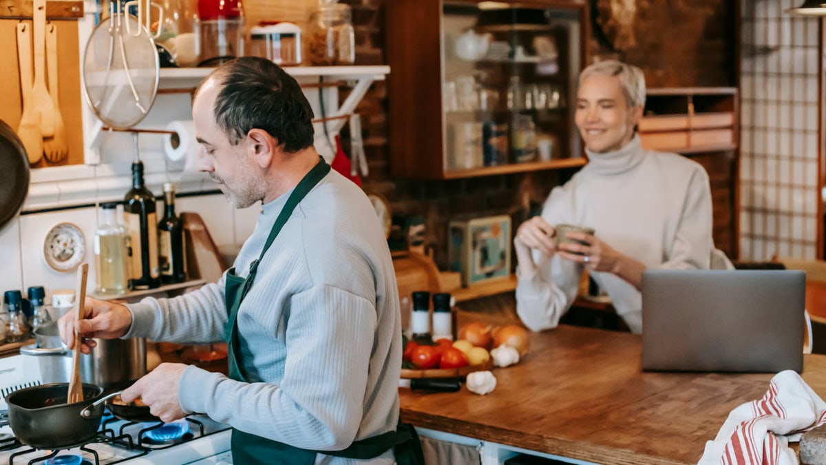 Focused male preparing dish on gas stove in kitchen with smiling wife having drink and using laptop